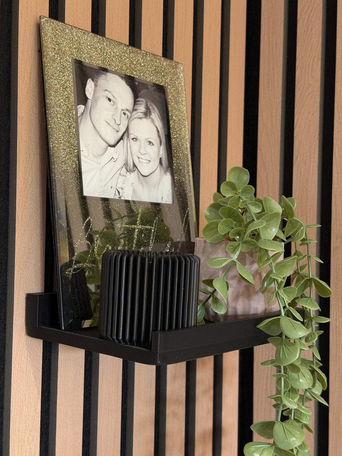 Close-up of a framed black-and-white photo resting on a picture ledge shelf with a trailing plant, mounted on a slat wall
