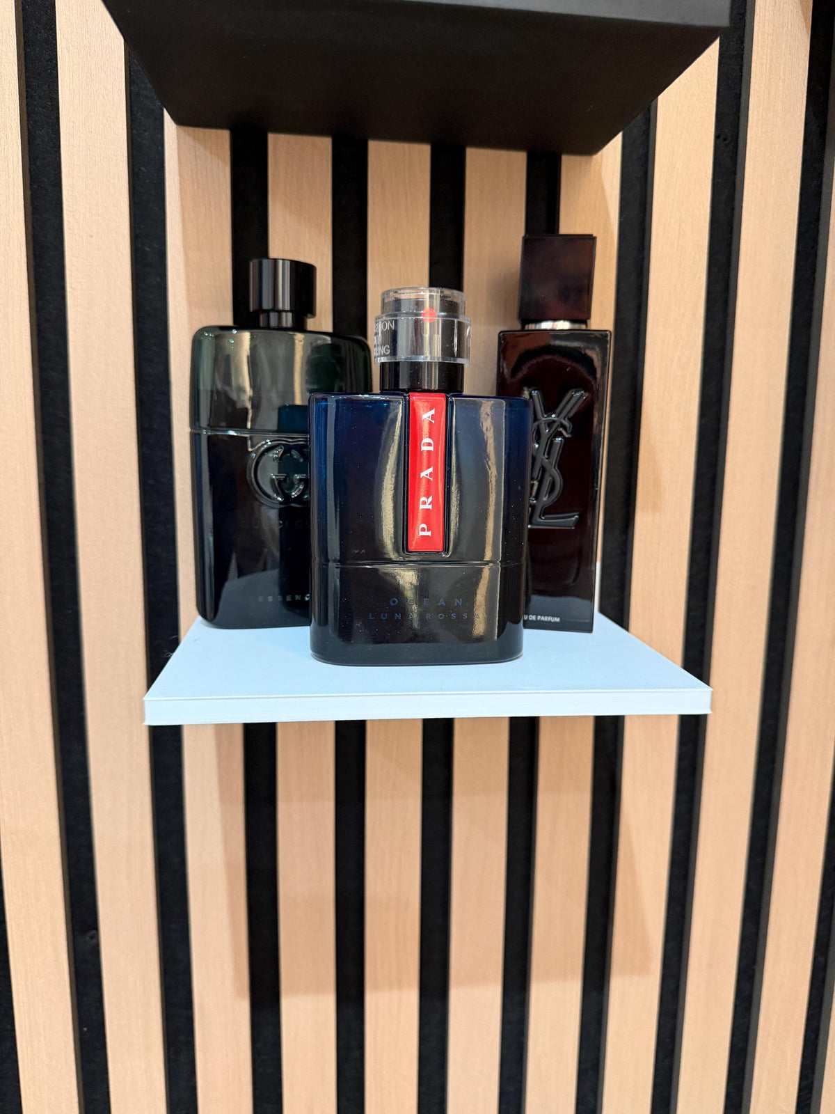White perfume shelf on a slat wall displaying dark fragrance bottles with plant in background