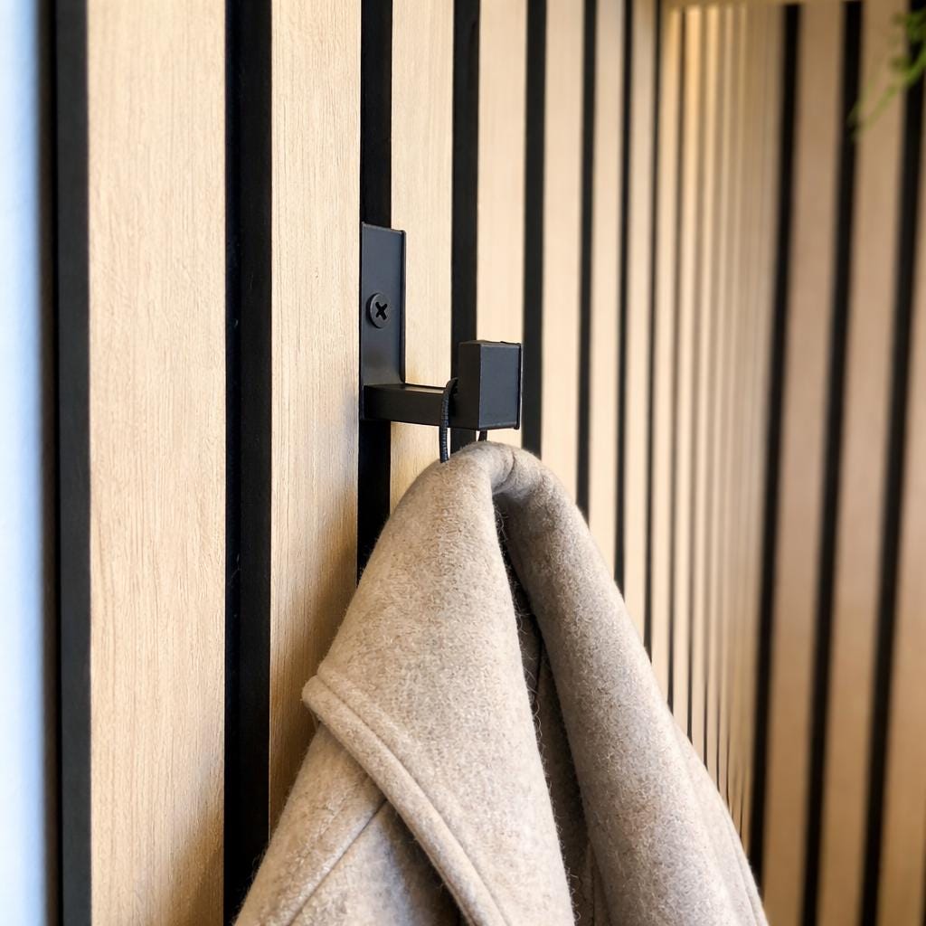 Matte black hook clipped onto an acoustic slat wall panel with a beige wool coat hanging from it